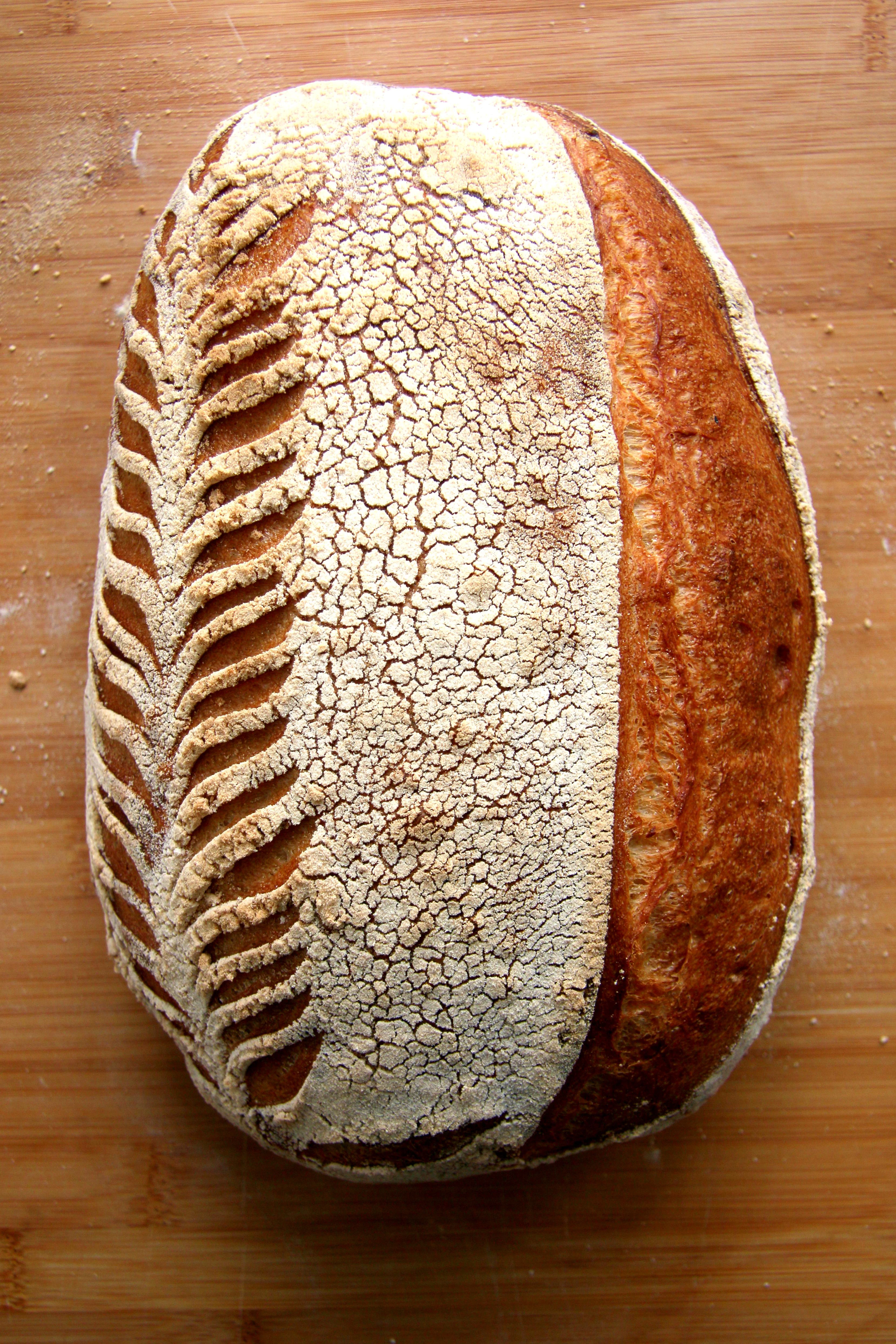 A rustic sourdough loaf with a wheat-stalk pattern scored into its floured crust, resting on a wooden board