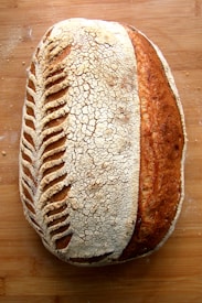 A beautifully crafted loaf of sourdough bread with intricate scoring patterns on the crust, lying on a wooden surface. The bread has a crackling texture with a blend of light and dark brown hues, indicating a well-baked exterior.