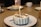 Several small round cakes with white frosting and chocolate drizzles are placed on white plates with brown rims. Each cake features a lit candle, and they are set on a wooden table. In the background, there are a few white cups and a basket.