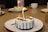 Several small round cakes with white frosting and chocolate drizzles are placed on white plates with brown rims. Each cake features a lit candle, and they are set on a wooden table. In the background, there are a few white cups and a basket.