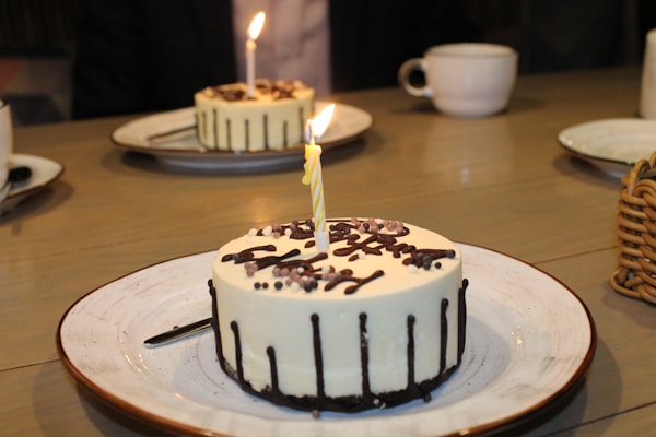 Several small round cakes with white frosting and chocolate drizzles are placed on white plates with brown rims. Each cake features a lit candle, and they are set on a wooden table. In the background, there are a few white cups and a basket.