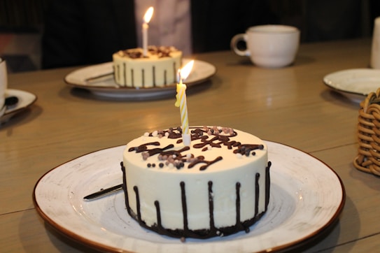 Several small round cakes with white frosting and chocolate drizzles are placed on white plates with brown rims. Each cake features a lit candle, and they are set on a wooden table. In the background, there are a few white cups and a basket.