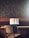 A wooden piano sits against a textured wall, with an open music book placed on the music stand. A simple chair with a fabric seat is positioned in front of the piano. The colors are earthy and muted, and a small cup is placed on top of the piano.