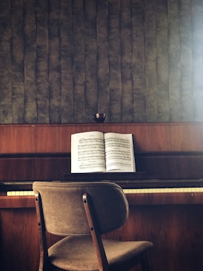 A wooden piano sits against a textured wall, with an open music book placed on the music stand. A simple chair with a fabric seat is positioned in front of the piano. The colors are earthy and muted, and a small cup is placed on top of the piano.