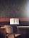 A wooden piano sits against a textured wall, with an open music book placed on the music stand. A simple chair with a fabric seat is positioned in front of the piano. The colors are earthy and muted, and a small cup is placed on top of the piano.