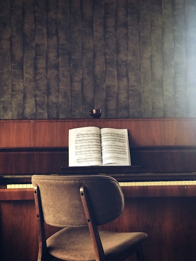 A wooden piano sits against a textured wall, with an open music book placed on the music stand. A simple chair with a fabric seat is positioned in front of the piano. The colors are earthy and muted, and a small cup is placed on top of the piano.