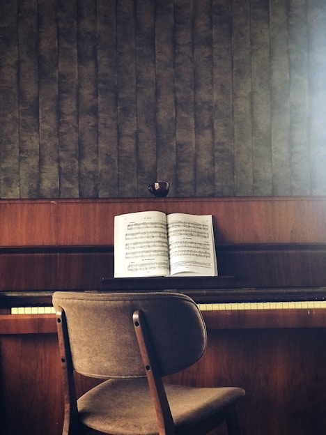 A wooden piano sits against a textured wall, with an open music book placed on the music stand. A simple chair with a fabric seat is positioned in front of the piano. The colors are earthy and muted, and a small cup is placed on top of the piano.