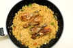 A black frying pan contains a serving of cooked pasta and noodles mixed with vegetables such as broccoli and bell peppers. Three grilled chicken drumsticks are placed on top of the pasta, creating a visually appealing dish.