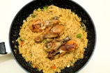 A black frying pan contains a serving of cooked pasta and noodles mixed with vegetables such as broccoli and bell peppers. Three grilled chicken drumsticks are placed on top of the pasta, creating a visually appealing dish.