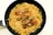 A black frying pan contains a serving of cooked pasta and noodles mixed with vegetables such as broccoli and bell peppers. Three grilled chicken drumsticks are placed on top of the pasta, creating a visually appealing dish.