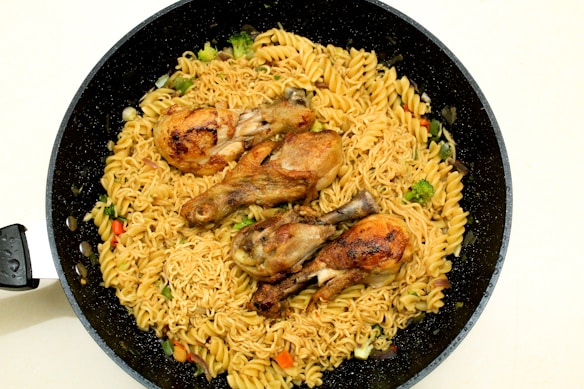A black frying pan contains a serving of cooked pasta and noodles mixed with vegetables such as broccoli and bell peppers. Three grilled chicken drumsticks are placed on top of the pasta, creating a visually appealing dish.