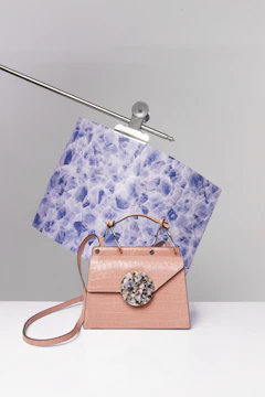 A stylish handbag with a pink crocodile texture sits on a white surface. It features a round, multicolored decorative piece on the front flap. Behind the handbag, a piece of blue and white patterned paper is clamped onto a metal rod.