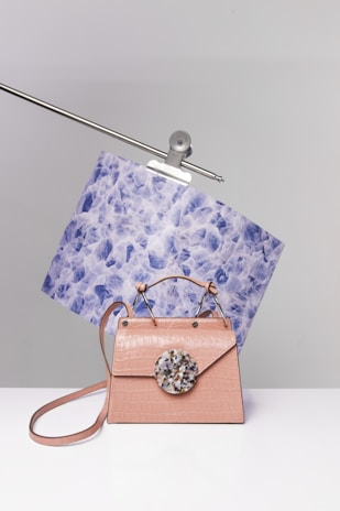 A stylish handbag with a pink crocodile texture sits on a white surface. It features a round, multicolored decorative piece on the front flap. Behind the handbag, a piece of blue and white patterned paper is clamped onto a metal rod.