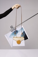A hand is holding a stylish handbag with an elongated brown strap. The handbag features a unique design with a white body and a beige top flap. Attached to it is a circular orange element. Behind the handbag, a piece of abstract art featuring blue and white colors is clipped to a metal rod.