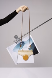A hand is holding a stylish handbag with an elongated brown strap. The handbag features a unique design with a white body and a beige top flap. Attached to it is a circular orange element. Behind the handbag, a piece of abstract art featuring blue and white colors is clipped to a metal rod.