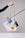 A hand is holding a stylish handbag with an elongated brown strap. The handbag features a unique design with a white body and a beige top flap. Attached to it is a circular orange element. Behind the handbag, a piece of abstract art featuring blue and white colors is clipped to a metal rod.