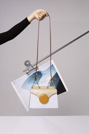 A hand is holding a stylish handbag with an elongated brown strap. The handbag features a unique design with a white body and a beige top flap. Attached to it is a circular orange element. Behind the handbag, a piece of abstract art featuring blue and white colors is clipped to a metal rod.