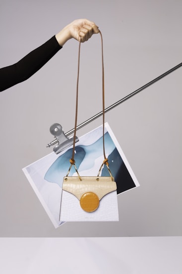 A hand is holding a stylish handbag with an elongated brown strap. The handbag features a unique design with a white body and a beige top flap. Attached to it is a circular orange element. Behind the handbag, a piece of abstract art featuring blue and white colors is clipped to a metal rod.