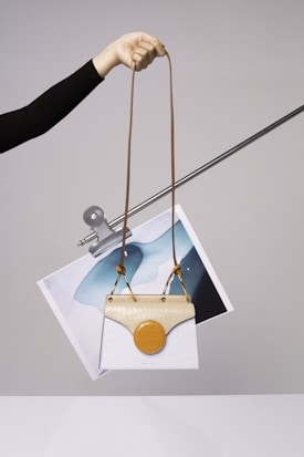 A hand is holding a stylish handbag with an elongated brown strap. The handbag features a unique design with a white body and a beige top flap. Attached to it is a circular orange element. Behind the handbag, a piece of abstract art featuring blue and white colors is clipped to a metal rod.