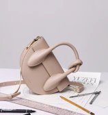 A stylish beige handbag with a unique sculptural design sits atop a table. Surrounding the bag are various drawing tools including a pencil, pens, a ruler, and a pair of compasses, all positioned on top of architectural sketches.