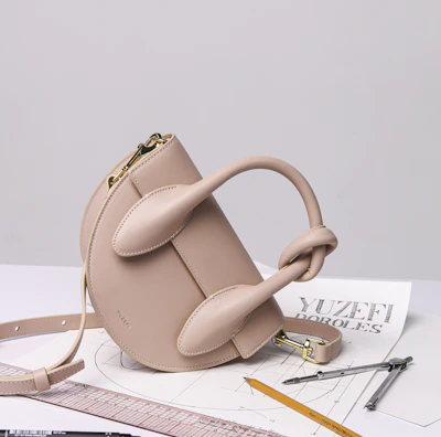 A stylish beige handbag with a unique sculptural design sits atop a table. Surrounding the bag are various drawing tools including a pencil, pens, a ruler, and a pair of compasses, all positioned on top of architectural sketches.