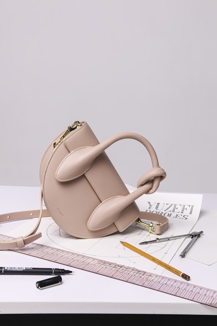 A stylish beige handbag with a unique sculptural design sits atop a table. Surrounding the bag are various drawing tools including a pencil, pens, a ruler, and a pair of compasses, all positioned on top of architectural sketches.