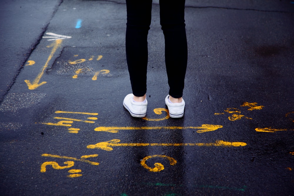 person in black pants and white sneakers standing on black asphalt road