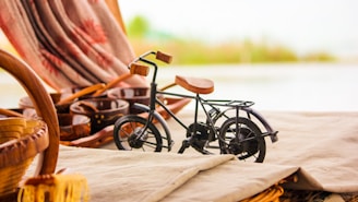 A small decorative bicycle made of black metal and wood sits on a beige fabric-covered surface. Surrounding the bicycle are wicker baskets and wooden objects, with a blurred background of a curtain featuring red and brown patterns.