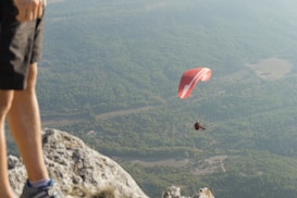 A red paraglider is soaring over a landscape filled with dense green forests and fields. The image is taken from a high vantage point on a rocky cliff, with a person's leg visible in the foreground, suggesting an adventurous viewpoint.