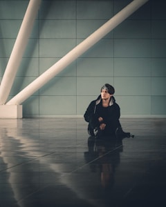 A person sitting on a glossy tiled floor, surrounded by a modern architectural background with large diagonal white beams against a muted teal wall. The person's relaxed posture and casual attire, including a hooded jacket and a headband, create a laid-back atmosphere.