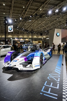 A sleek, futuristic racing car is displayed in a well-lit exhibition space. The car features a combination of white, blue, and purple colors with various sponsor logos. People are gathered around, capturing photos and admiring the vehicle. The setting includes a ceiling with a lattice of beams and spotlights, along with branding from a prominent car manufacturer.