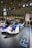 A sleek, futuristic racing car is displayed in a well-lit exhibition space. The car features a combination of white, blue, and purple colors with various sponsor logos. People are gathered around, capturing photos and admiring the vehicle. The setting includes a ceiling with a lattice of beams and spotlights, along with branding from a prominent car manufacturer.
