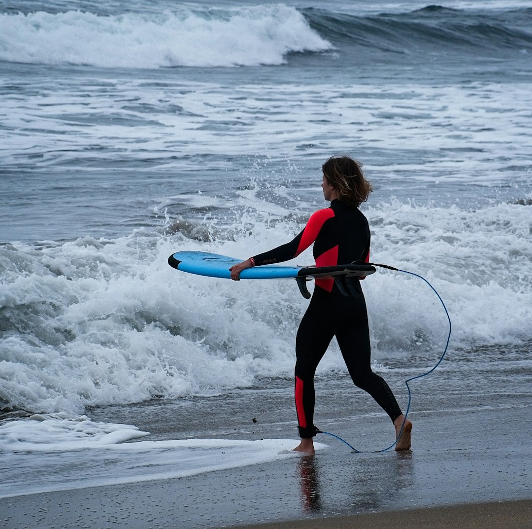 woman in black and red jacket holding blue surfboard on beach during daytime