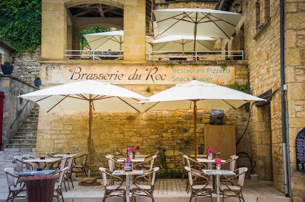 Outdoor seating area of a rustic restaurant with stone walls and large white umbrellas providing shade. There are several tables and chairs with pink flowers on the tables. A sign on the wall reads 'Brasserie du Roc'.