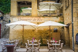 Outdoor seating area of a rustic restaurant with stone walls and large white umbrellas providing shade. There are several tables and chairs with pink flowers on the tables. A sign on the wall reads 'Brasserie du Roc'.