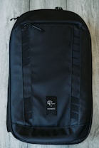A sleek, black backpack with a modern design rests on a wooden floor. It features textured fabric, multiple zippers, and a logo in the center.