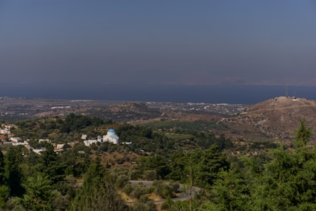 A serene landscape featuring a coastal town with a notable blue-domed building amidst lush greenery. The horizon shows a vast expanse of water with distant mountains under a clear sky. Rolling hills with sparse vegetation add depth to the scenery.