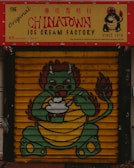 A colorful storefront features a signage for The Original Chinatown Ice Cream Factory with red and yellow colors. Below the sign, a painted shutter displays a cartoon dragon holding an ice cream bowl. The dragon has a green body, red accents, and appears friendly and playful.