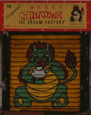 A colorful storefront features a signage for The Original Chinatown Ice Cream Factory with red and yellow colors. Below the sign, a painted shutter displays a cartoon dragon holding an ice cream bowl. The dragon has a green body, red accents, and appears friendly and playful.