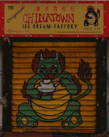 A colorful storefront features a signage for The Original Chinatown Ice Cream Factory with red and yellow colors. Below the sign, a painted shutter displays a cartoon dragon holding an ice cream bowl. The dragon has a green body, red accents, and appears friendly and playful.