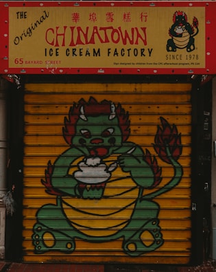 A colorful storefront features a signage for The Original Chinatown Ice Cream Factory with red and yellow colors. Below the sign, a painted shutter displays a cartoon dragon holding an ice cream bowl. The dragon has a green body, red accents, and appears friendly and playful.
