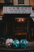 A shopfront with a sign reading 'New Mee Lun Co.' in both English and Chinese characters. Below the sign, three bags of garbage are placed on the sidewalk, filled with plastic containers and cans. The scene is dimly lit, with a neon sign visible through a metal gate.