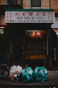 A shopfront with a sign reading 'New Mee Lun Co.' in both English and Chinese characters. Below the sign, three bags of garbage are placed on the sidewalk, filled with plastic containers and cans. The scene is dimly lit, with a neon sign visible through a metal gate.