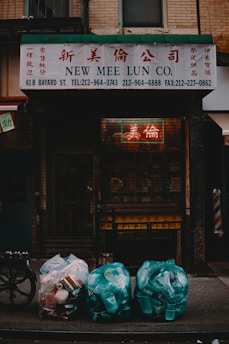 A shopfront with a sign reading 'New Mee Lun Co.' in both English and Chinese characters. Below the sign, three bags of garbage are placed on the sidewalk, filled with plastic containers and cans. The scene is dimly lit, with a neon sign visible through a metal gate.