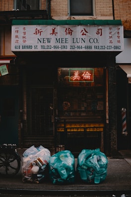 A shopfront with a sign reading 'New Mee Lun Co.' in both English and Chinese characters. Below the sign, three bags of garbage are placed on the sidewalk, filled with plastic containers and cans. The scene is dimly lit, with a neon sign visible through a metal gate.
