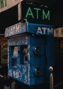 An old, worn ATM machine with a peeling blue exterior is positioned in a dimly lit setting. The illuminated sign on top displays the letters 'ATM' in green. The machine's appearance suggests it has been in use for a long time.