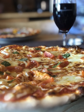 A close-up of a freshly baked pizza topped with melted cheese, tomato slices, and fresh basil leaves. In the background, there is a dark glass of red wine, slightly out of focus, on a wooden table.
