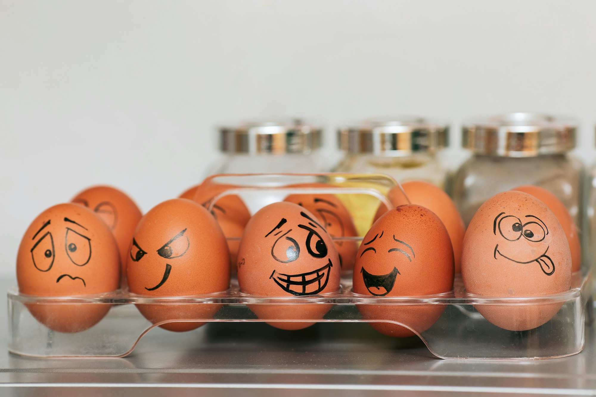 A photograph (wallpaper, picture) of eggs with different emotions against a background of blurred spices and a smooth gray wall.  Whole painted eggs in the kitchen, expressing a spectrum of emotions from sadness to joy or madness. Eggs with smile and crazy face.