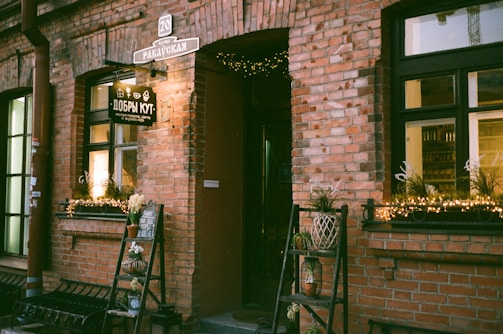 A charming storefront with a rustic brick exterior. There are two large windows adorned with fairy lights and potted plants sitting on window sills. An open wooden door leads inside, while a small chalkboard sign and a decorative plant stand are positioned by the entrance. The sign on the shop reads in a foreign language, adding a cozy, inviting atmosphere.