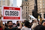 A large crowd of people gathers in a city setting, holding various signs. One prominent sign in the forefront reads 'Sorry, We're Closed Due to Climate Crisis #ClimateStrike.' The background features tall buildings and a black sculpture, suggesting an urban environment.